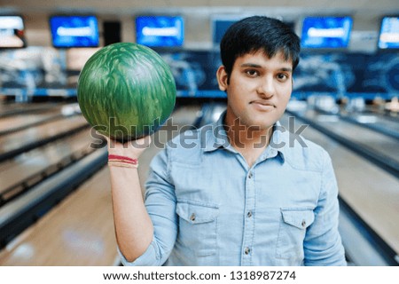 Stylish asian man in jeans shirt standing at bowling alley with ball at hand.