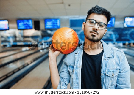 Stylish beard asian man in jeans jacket and glasses standing at bowling alley with ball at hand.