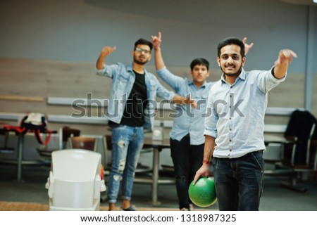 South asian man in jeans shirt standing at bowling alley with ball on hands. Guy is preparing for a throw. Friends support him loudly.