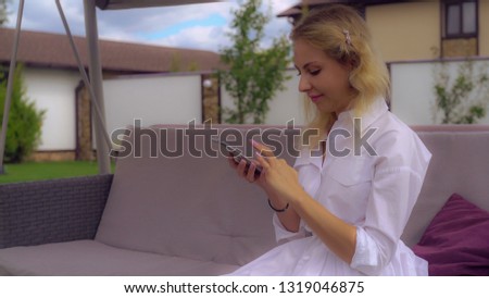 Woman looking at new smartphone from different side. Female holding mobile phone. Businesswoman using cell sits on the traditional english porch swing in the garden in backyard near the house
