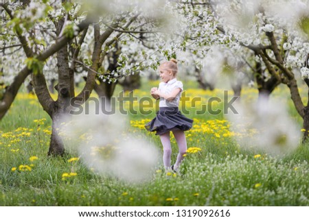 Blonde happy child girl in the white blouse and gray skirt dancing in the blossom cherry garden. Spring season. Trees with flowers on the background.