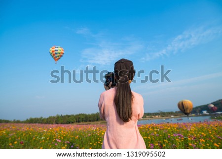 Beautiful asian woman watching colorful hot air balloons flying over field of flowers and take photo of balloon