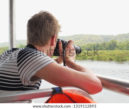 Young man taking a picture of the green landscape while sitting in a passenger boat.
