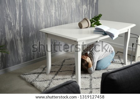 Young woman hiding under table during earthquake Royalty-Free Stock Photo #1319127458