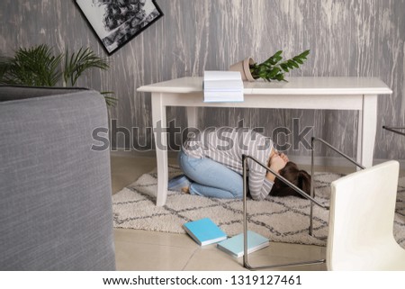 Young woman hiding under table during earthquake Royalty-Free Stock Photo #1319127461