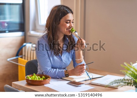 Happy young business woman enjoying a healthy salad at work. Young business woman eating salad at home, having healthy lunch at workplace while working on laptop 