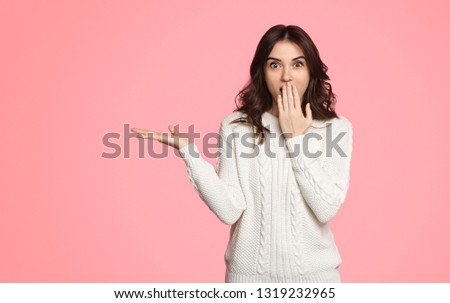Beautiful young lady in white sweater covering mouth with hand and presenting empty space on pink background