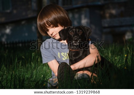 A boy with long hair is sitting in the grass. He is hugging a small funny black dog.