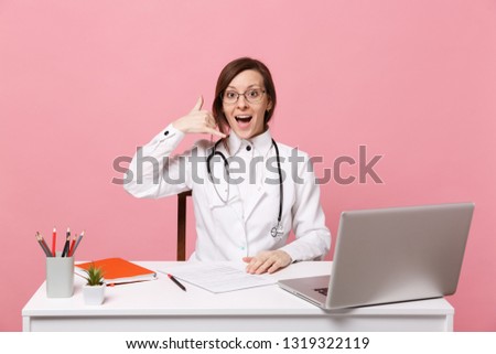 Beautiful female doctor sits at desk works on computer with medical document in hospital isolated on pastel pink background. Woman in medical gown glasses stethoscope. Healthcare medicine concept
