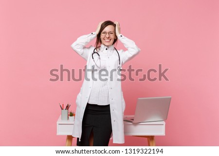 Cute female doctor stand in front of desk with pc computer, medical document in hospital isolated on pastel pink background. Woman in medical gown glasses stethoscope. Healthcare medicine concept