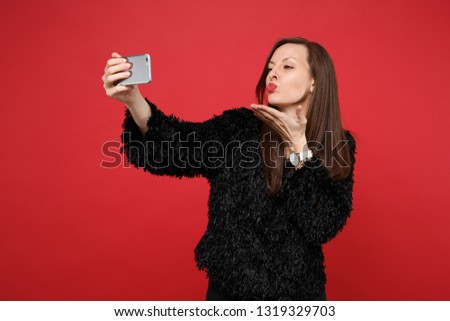 Pretty young girl in black fur sweater blowing sending air kiss doing selfie shot on mobile phone isolated on bright red wall background. People sincere emotions lifestyle concept. Mock up copy space