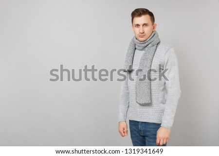 Portrait of smiling young man in gray sweater, scarf standing isolated on grey wall background in studio. Healthy fashion lifestyle, people sincere emotions, cold season concept. Mock up copy space