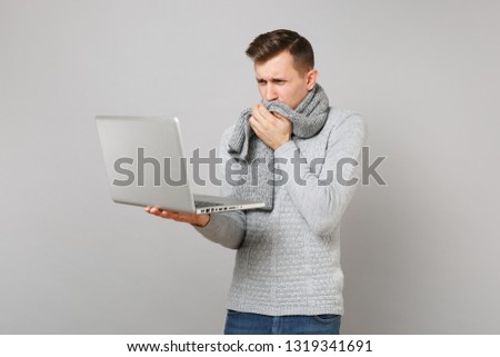 Coughing man in gray sweater covering mouth with scarf working on laptop pc computer isolated on grey background. Healthy lifestyle online treatment consulting cold season concept. Mock up copy space
