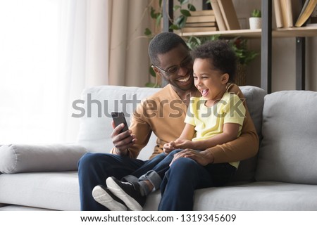 Happy african dad and toddler child son laughing looking at cellphone using funny smartphone mobile face app sitting on sofa, black father holding phone taking selfie with cute little kid boy at home Royalty-Free Stock Photo #1319345609