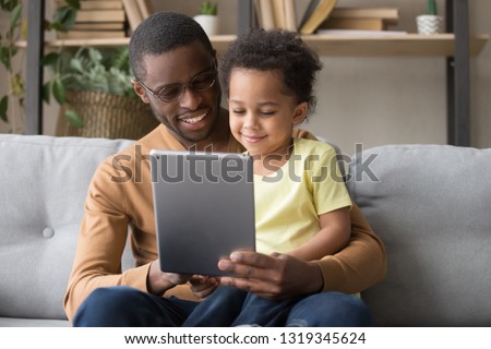 Happy african dad and little toddler son holding digital tablet doing online shopping at home, black father teaching kid boy use computer, daddy and child having fun playing app game sitting on sofa