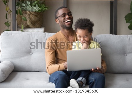 Happy black dad watching funny cartoons movie with little toddler son sitting on couch, african father and cute small kid boy having fun with computer, daddy and child laughing using laptop at home