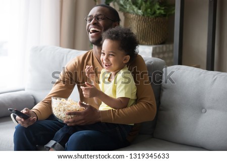Happy african american dad and kid son laughing holding popcorn remote control watching funny comedy movie tv show sitting on sofa, black father having fun with child boy viewing television at home Royalty-Free Stock Photo #1319345633