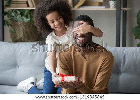 Cute african kid daughter closing eyes congratulating black dad with happy birthday giving gift box sitting on sofa, child girl making surprise preparing present on fathers day having fun with daddy