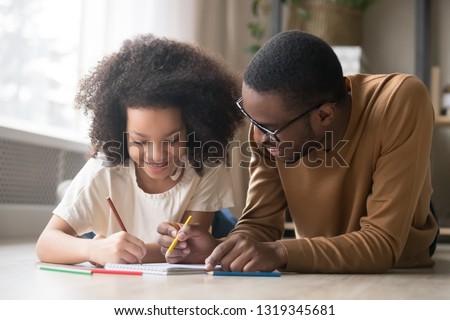 Caring black dad babysitter drawing with colored pencils teaching child girl lying on floor, african father enjoy helping kid daughter playing doing homework at home, happy family creative activity Royalty-Free Stock Photo #1319345681