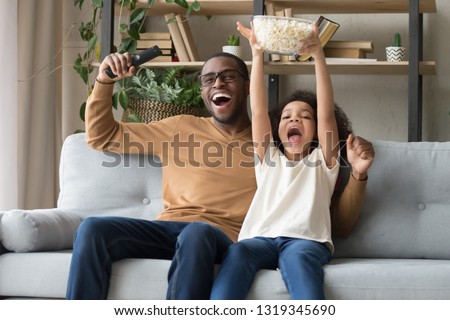 Happy overjoyed black family father with kid daughter sport fans watching tv game supporting football team, excited african dad and child girl celebrating victory soccer goal at home sitting on sofa Royalty-Free Stock Photo #1319345690