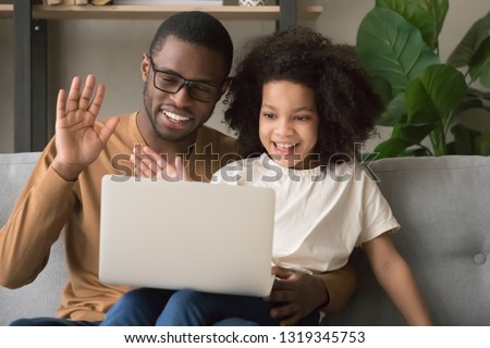 Happy family black dad with kid daughter waving hands making distance video call looking at laptop, african father and child girl looking talking to webcamera chatting online by computer webcam Royalty-Free Stock Photo #1319345753
