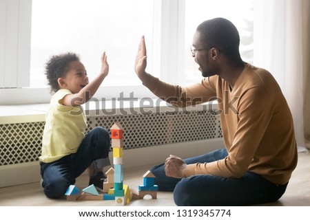 Happy black dad and cute toddler son giving high five playing with wooden blocks on floor at home, little kid boy enjoying game with african father, daddy child clapping hands having fun together Royalty-Free Stock Photo #1319345774