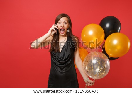 Excited young woman in little black dress holding air balloons, talking on mobile phone, screaming isolated on bright red background. Women's Day, Happy New Year birthday mockup holiday party concept