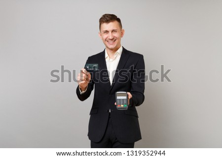 Joyful business man holding wireless modern bank payment terminal to process and acquire credit card payments, black card isolated on grey wall background. Achievement career wealth business concept