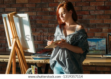 Artist painting. Studio workspace. Redhead female with easel mixing colors on wooden palette.