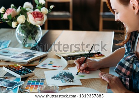Watercolor painting. Artist at work. Smiling female painter doing color mix brushstrokes. Sketches and palette supplies around.