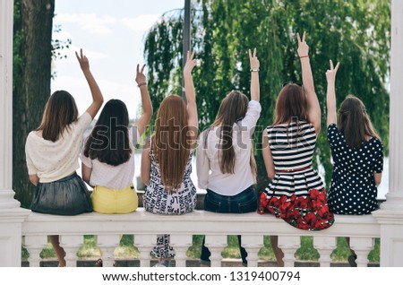 Girls girlfriends sit on the railing and raise one hand to up. They show a victory sign. Back view