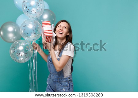 Laughing young girl hold red striped present box with gift ribbon celebrating with colorful air balloons isolated on blue turquoise background. Valentine's Day, Women's Day, birthday, holiday concept