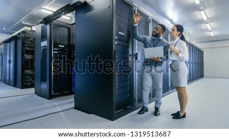 Black IT Technician with a Laptop Computer Gives a Tour to a Young Intern. They Talk in Data Center while Walking Next to Server Racks. Running Diagnostics or Doing Maintenance Work.
