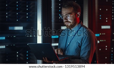 Bearded IT Specialist in Glasses is Working on Laptop in Data Center while Standing Near Server Rack. Running Diagnostics, Doing Maintenance Work. Emergency Red Light from Side Illuminating Specialist Royalty-Free Stock Photo #1319513894