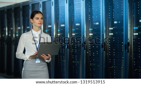 Beautiful Data Center Female IT Technician Walking Through Server Rack Corridor with a Laptop Computer. She is Visually Inspecting Working Server Cabinets. Royalty-Free Stock Photo #1319513915