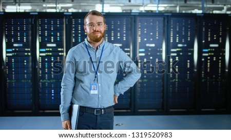 Bearded IT Specialist in Glasses is Standing in Data Center Next to Server Racks, Smiling to the Camera and Holding Laptop. Running Diagnostics or Doing Maintenance Work.