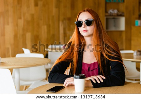 Beautiful redhead model with sunglasses in cafeteria. Cellphone and coffee cup on the table with blurred background.