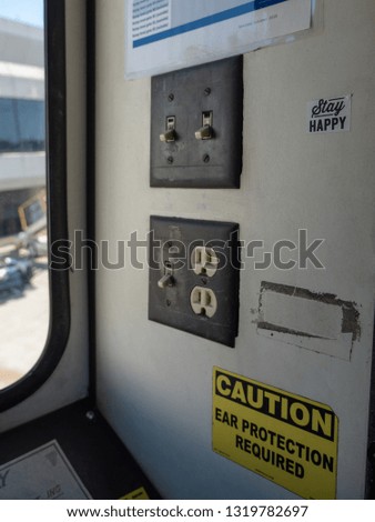 A "Stay Happy" sticker near light switches and outlets on the wall of a jet bridge near a yellow "Caution: Ear Protection Required" sign