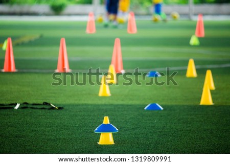 Cone markers is soccer training equipment on green artificial turf with blurry kid players training background. Material for training class of football academy