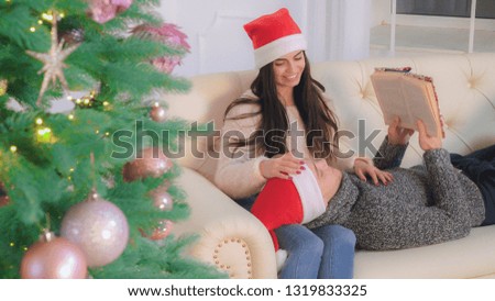 woman and man spend winter holidays traditional at home. Resting on the sofa reading book talking. Young attractive woman smiling and looking with love on her boyfriend. Couple relaxing near christmas