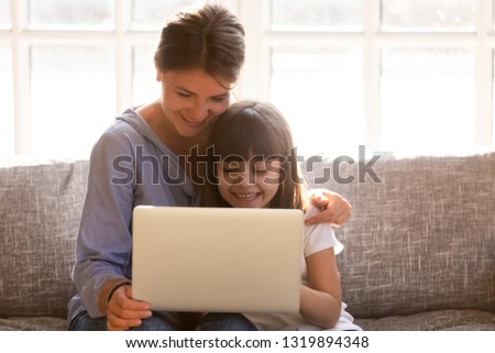 Happy mom with little child daughter having fun online with laptop computer on couch, mother and kid girl laughing watching cartoons or making call doing internet shopping using applications together