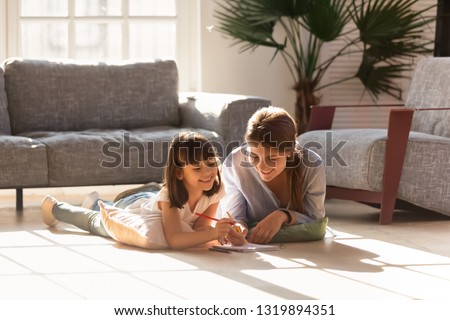 Happy mom helping child daughter drawing with colored pencils laying on warm floor together, smiling baby sitter mother teaching cute kid learning creative activity play laugh at home in living room