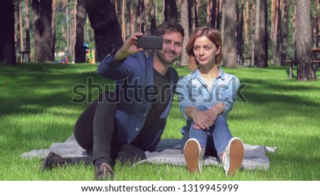 Tired woman and smiling man sitting on the grass take selfie photo at the park. Beautiful couple in love have fun on a date. Casual girlfriend and boyfriend in sunny summer day. Happy people wearing