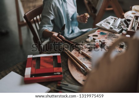 Close up of the table in art studio covered with painting equipment and female artist taking paintbrushes and spatula to her hand