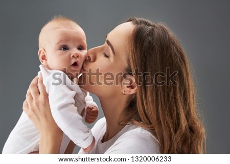 Close up photo of pretty baby looking aside with open mouth while mum kissing he in cheek isolated on gray background