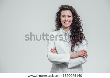 Portrait of happy young smiling girl doctor. Dressed in a white robe. Evenly standing with crossed hands on a gray background. Royalty-Free Stock Photo #1320096140