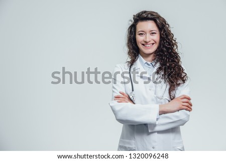 Portrait of happy young smiling girl doctor. Dressed in a white robe. Evenly standing with crossed hands on a gray background. Royalty-Free Stock Photo #1320096248