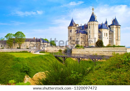 Front facade, wooden bridge, and entrance to majestic Château de Saumur castle and surrounding walls in France. Warm spring morning, vibrant blue sky with dramatic clouds, green lawn, calm atmosphere Royalty-Free Stock Photo #1320097472