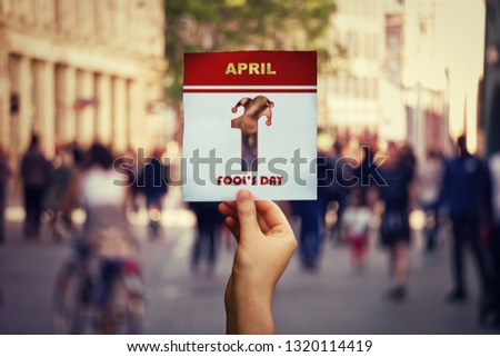 Hand holding a calendar page with 1 april fool's day symbol over crowded street background. International fun holiday.