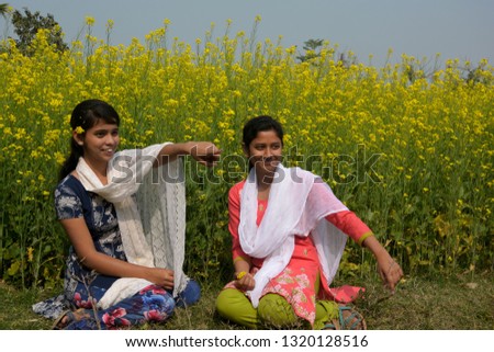 Close up two beautiful girls sitting in a rapeseed, mustard flower field with two flowers on their ears smiling, talking and pointing  something, conceptual picture, selective focusing